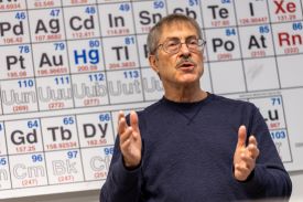 A man with a mustache, blue sweater and eyeglasses stands with his hands raised at chest height while speaking in front of a periodic table of elements.
