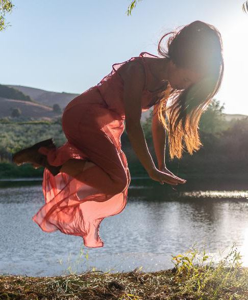 dancer in rose colored dress jumps in air outdoors with body of water & mountains in background