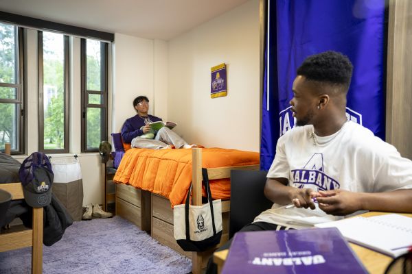 Two young men sit in a dormatory room, one talking from a desk with notebooks on it, the other listening from his bed near the windows.