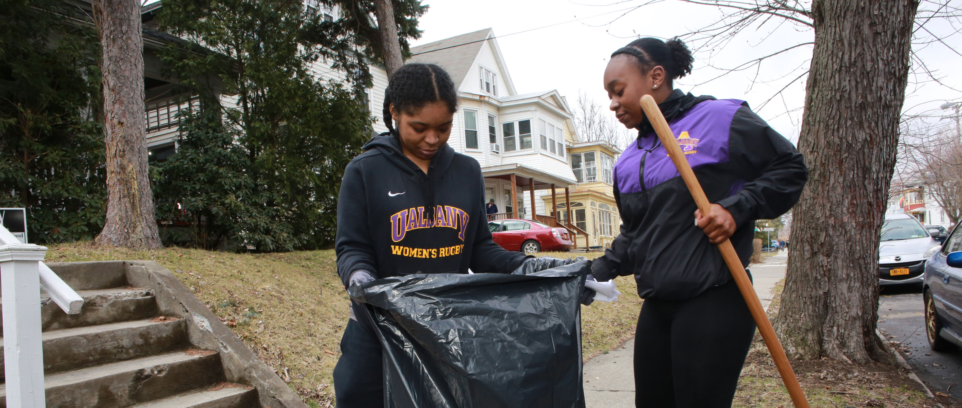 Two students hold a black trash bag open as they participate in a Pine Hills neighborhood clean-up.