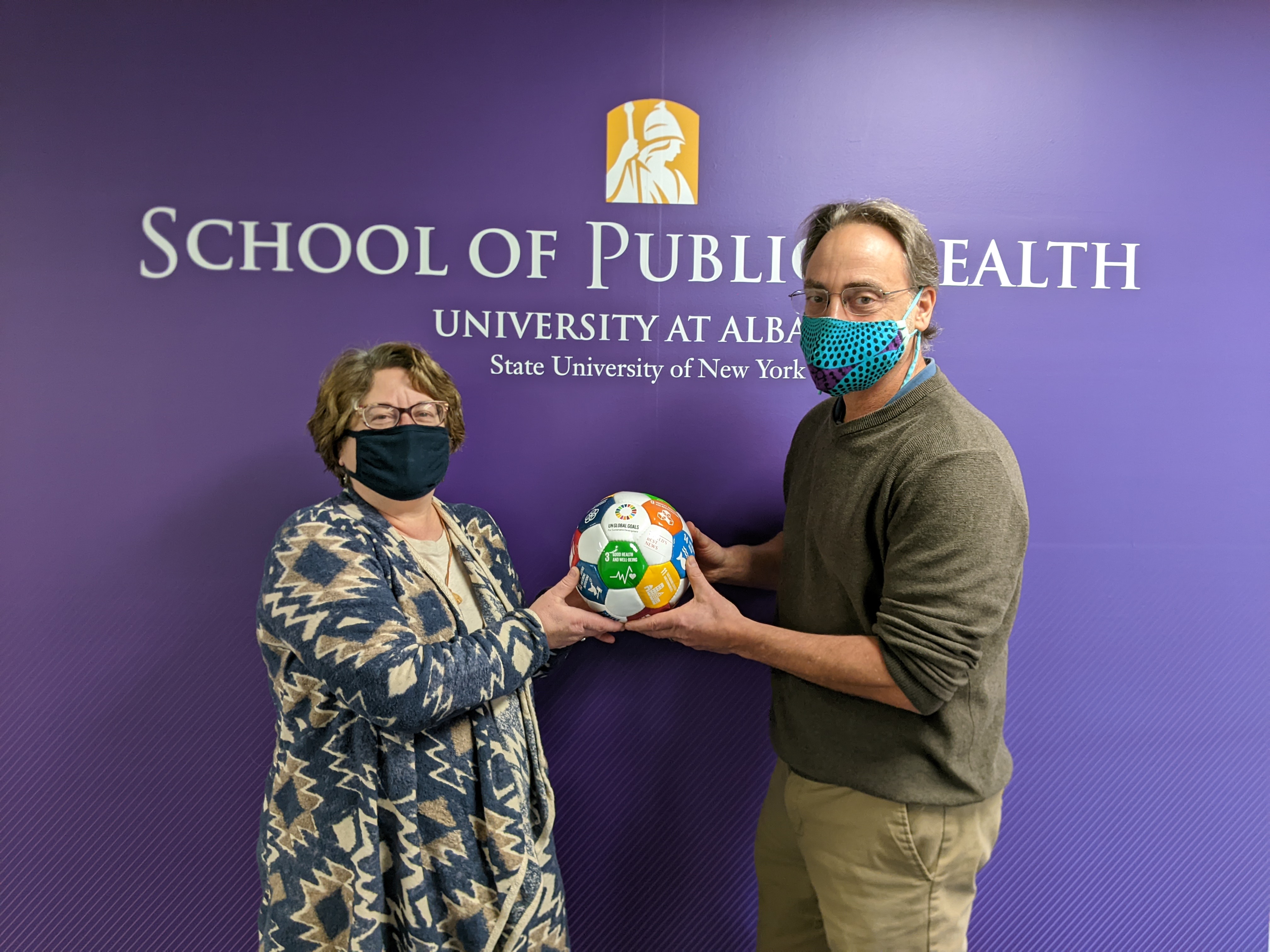 John and Lynne stand in front of a purple wall that says "The School of Public Health" in white text. They are wearing face masks and are holding a soccer ball from the UN that has the Sustainability Goals written on it.