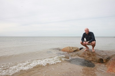 A man squats on a rock along a sandy coastline on an overcast day