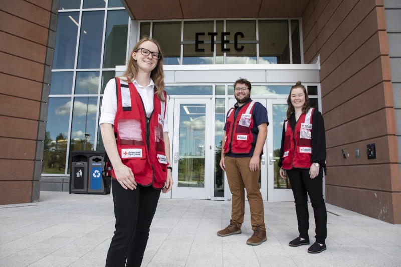 Three students wearing American Red Cross volunteer vests pose for a picture outside of the entrance to a building. The letters “E-T-E-C” are above the building entrance.