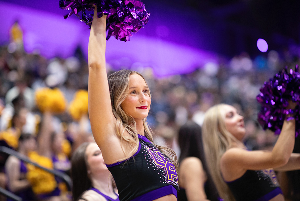 A Ualbany Cheerleader smiles, raising a pom pom into the air at an event.