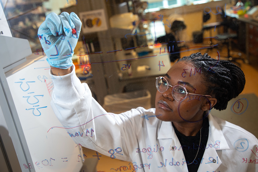 A person wearing a lab coat write equations on a glass board in marker, they wear an expression of concentration.