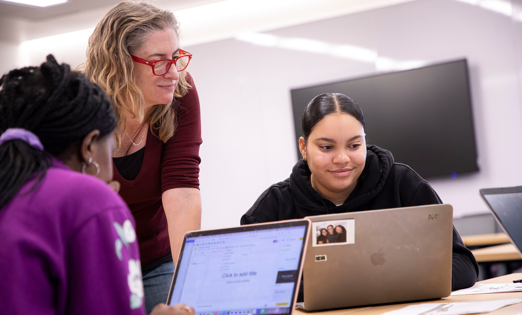 An instructor smiles as she leans over a table and speaks with two students working on laptops.