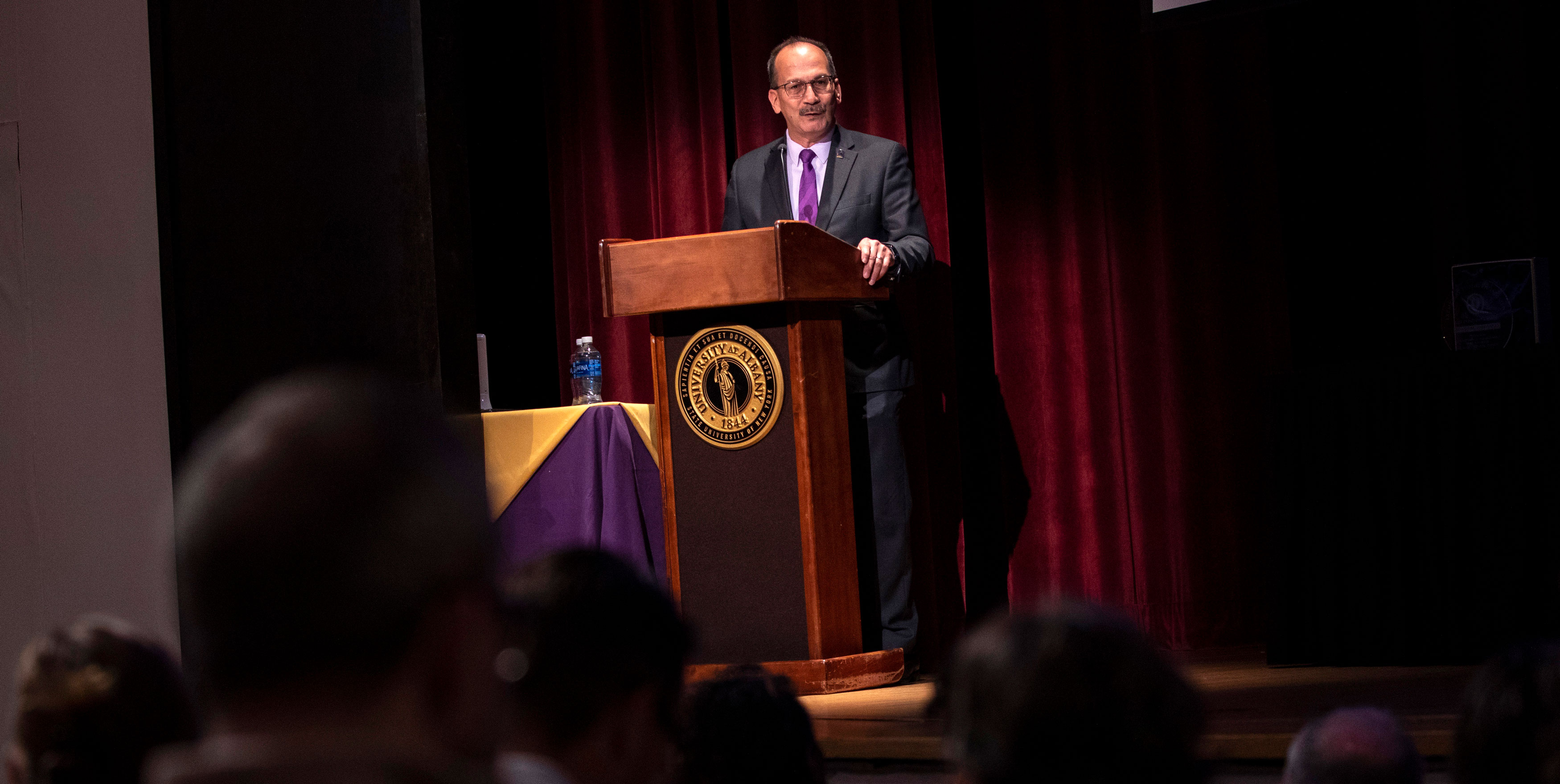 UAlbany President Havidán Rodríguez speaks from behind a podium with the UAlbany seal to a gathered audience inside an auditorium.