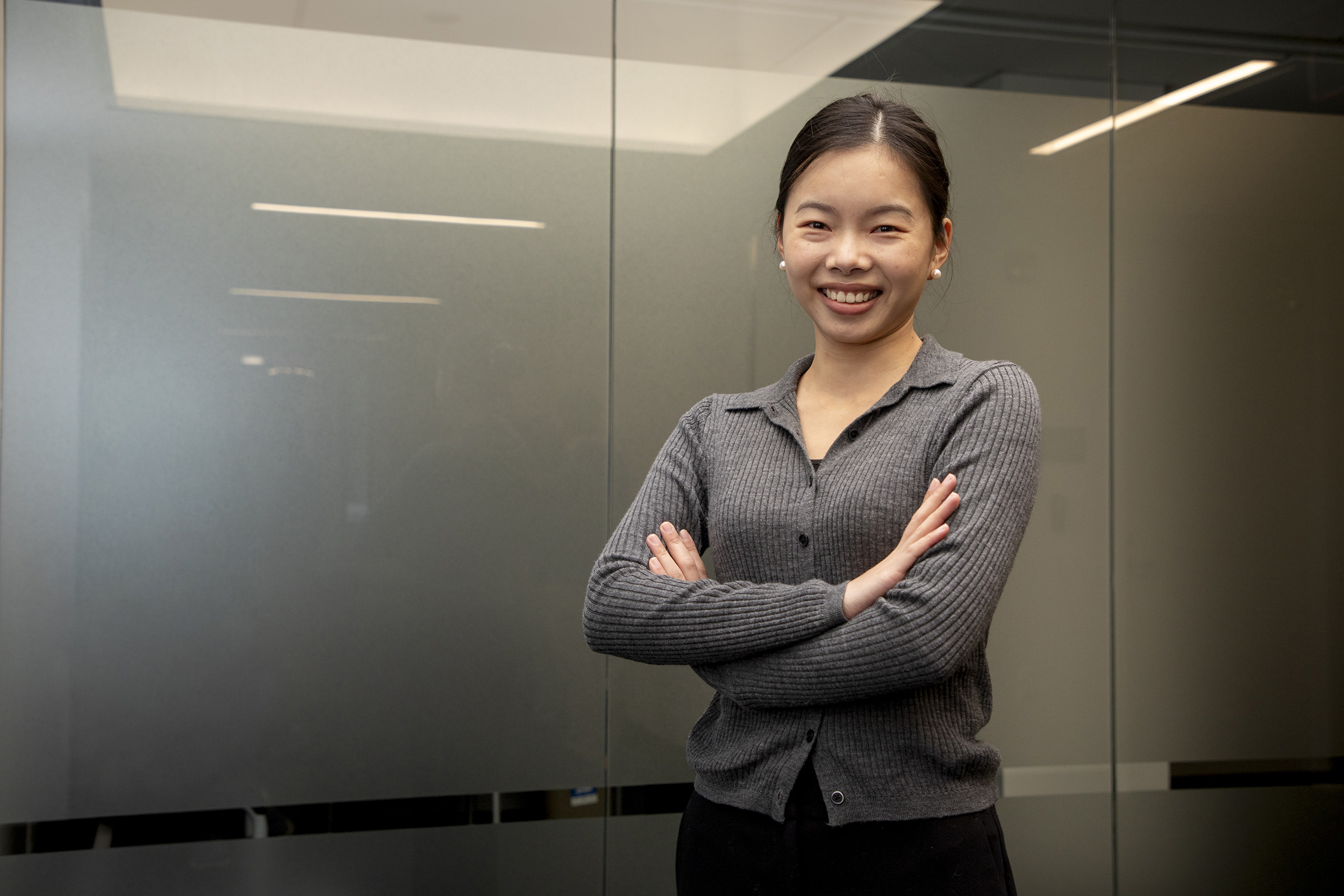 A girl with dark hair and white earings smiles as she stands in front of a frosted glass panel