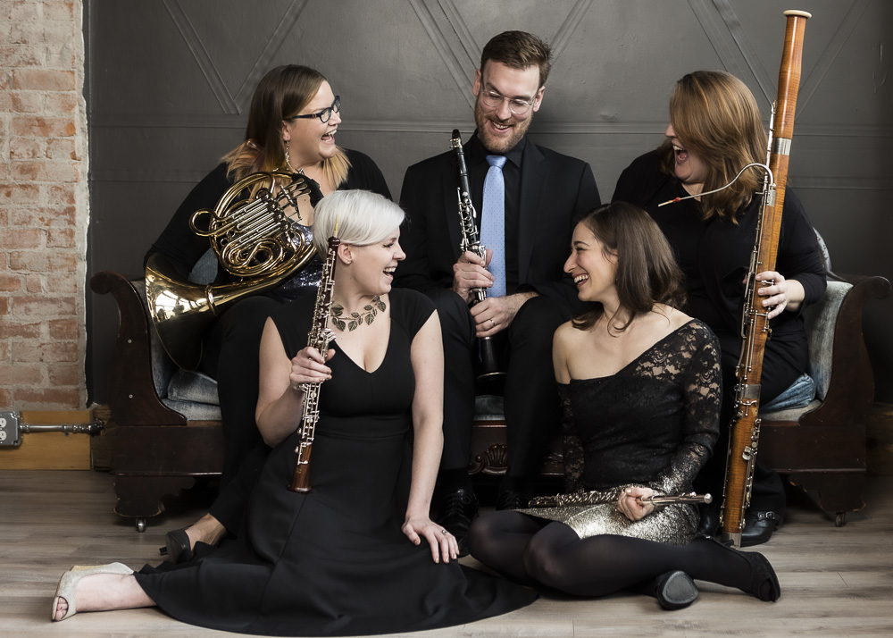 five musicians with instruments sit in a clump and smile at each other