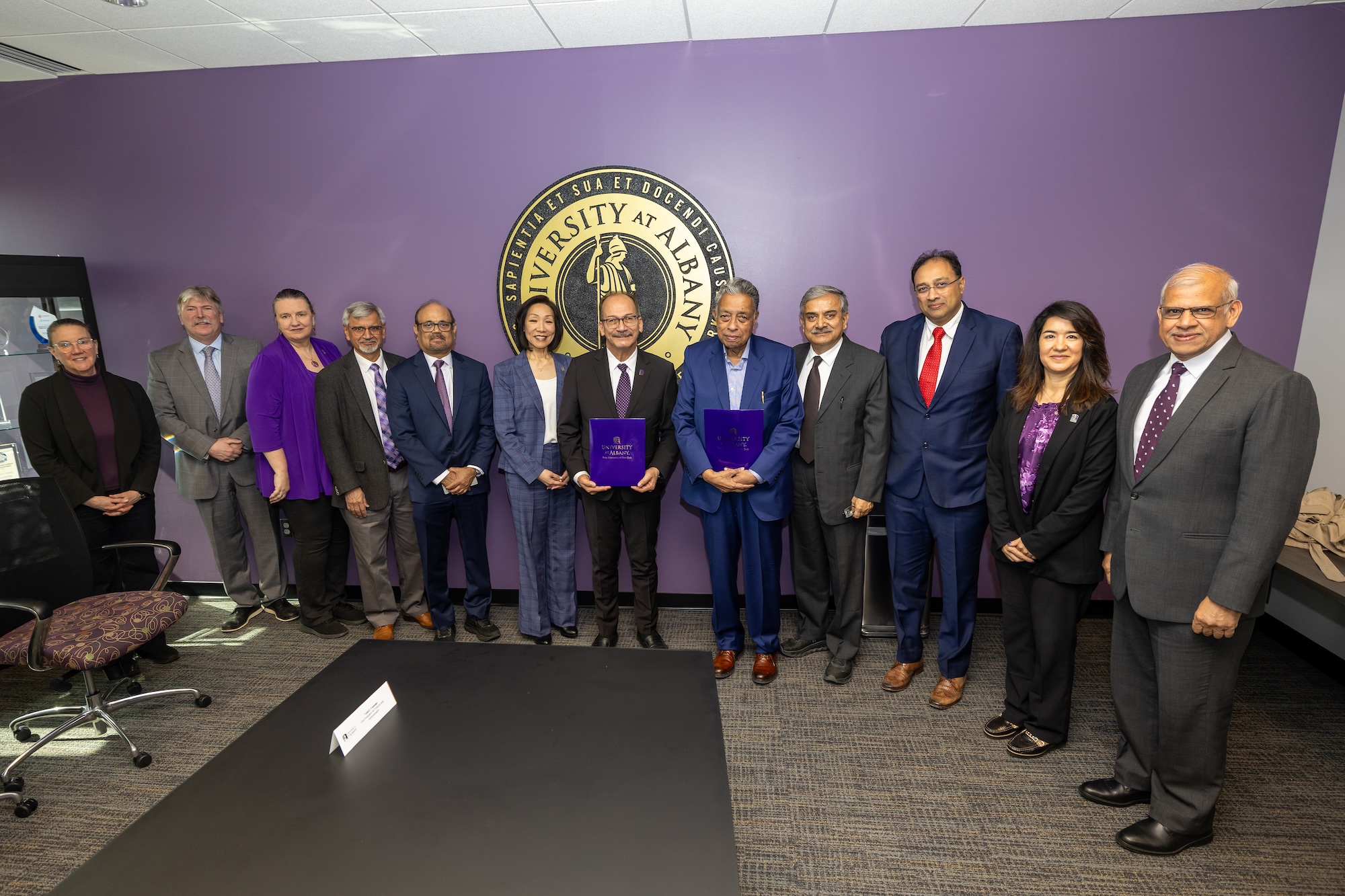 People line up for a group photo against a purple wall with a UAlbany seal