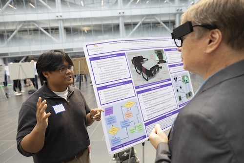 A man wearing a pair of smart glasses talks to a younger man standing in front of a poster.