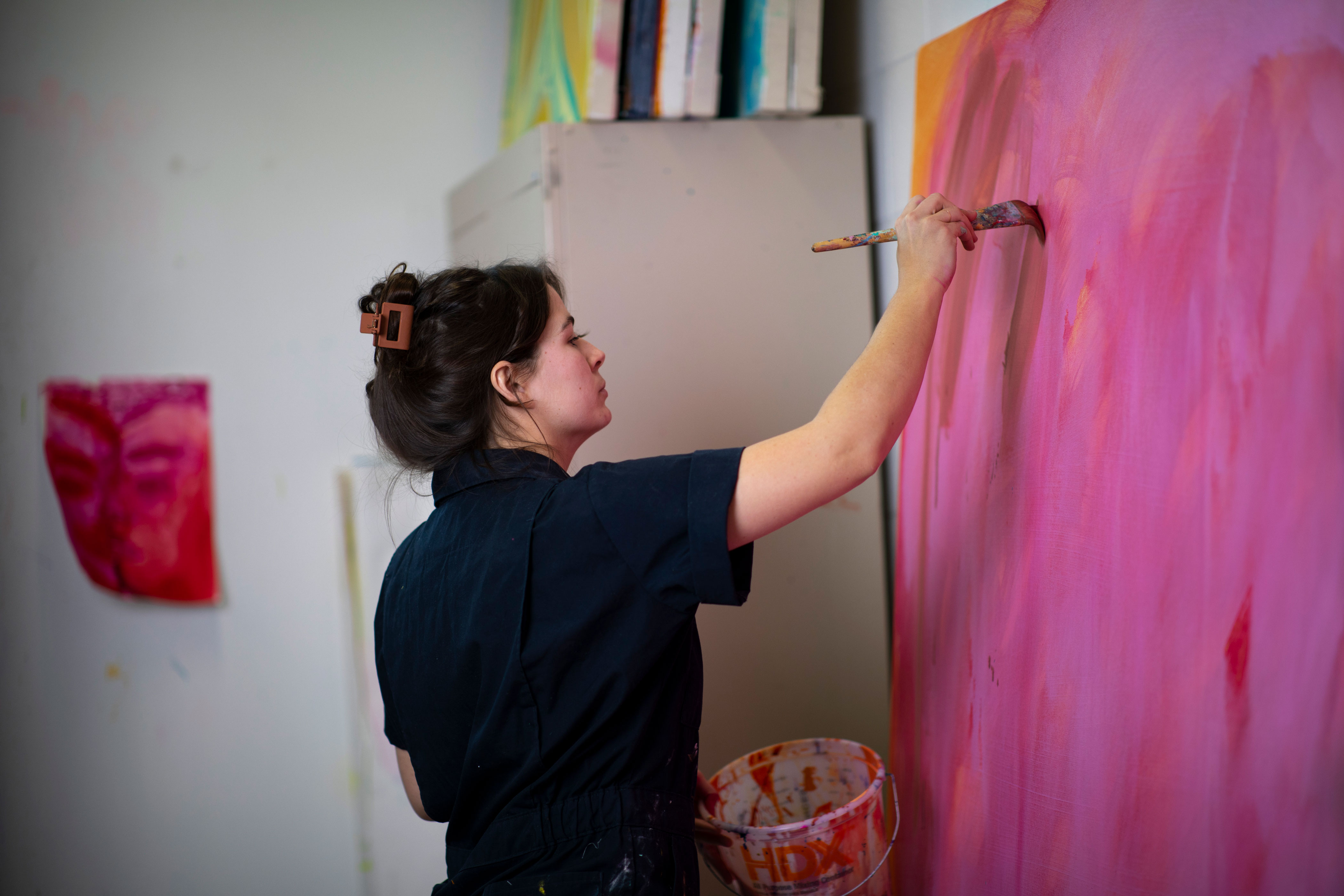 A student in navy coveralls, with her hair clipped back, uses a brush on a pink abstract painting.