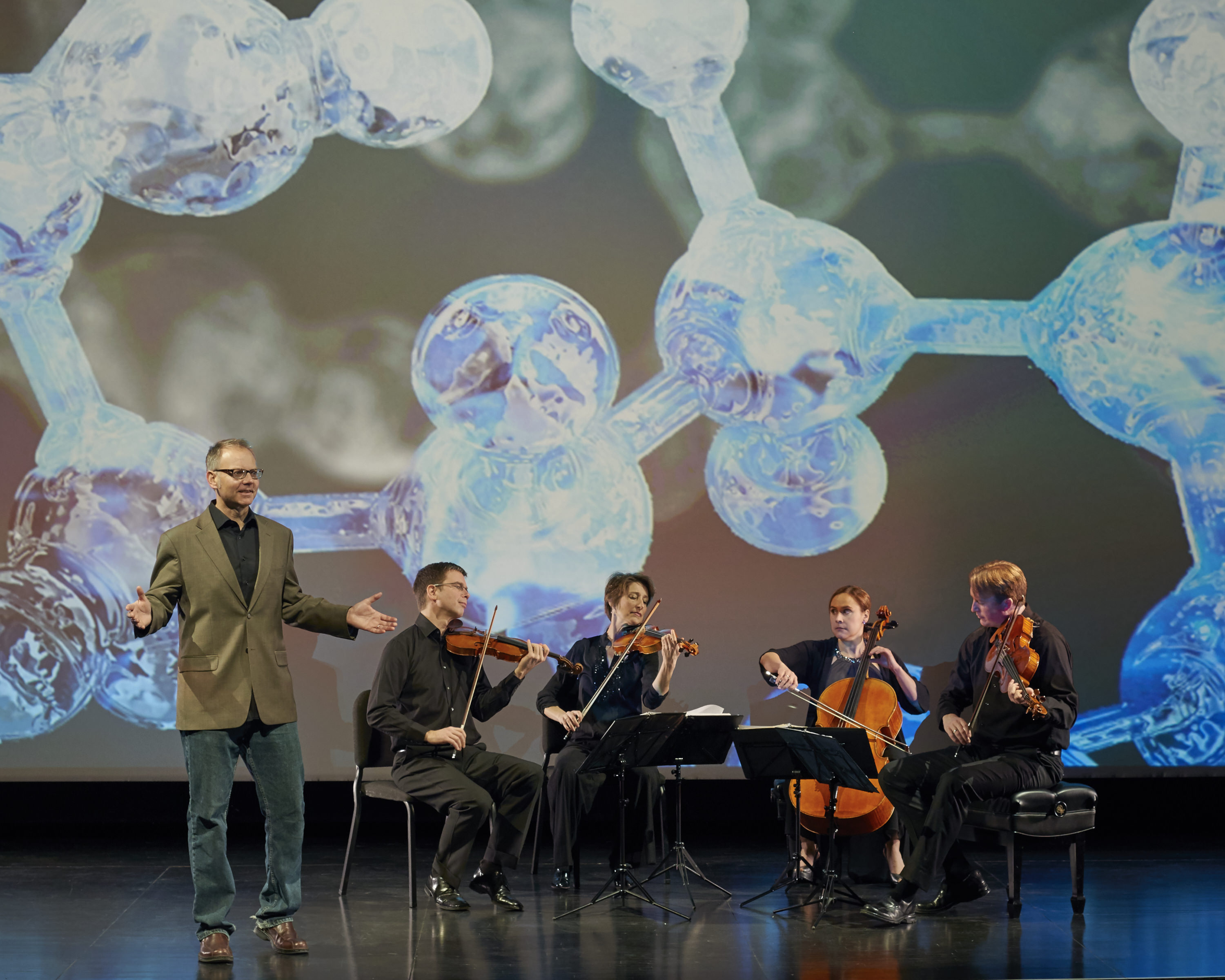 performer stands speaking by seated musicians playing instruments with image of molecular structures in background