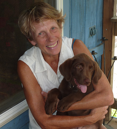 A woman with short strawberry blonde hair holds a chocolate-colored puppy in her arms, its tongue wagging
