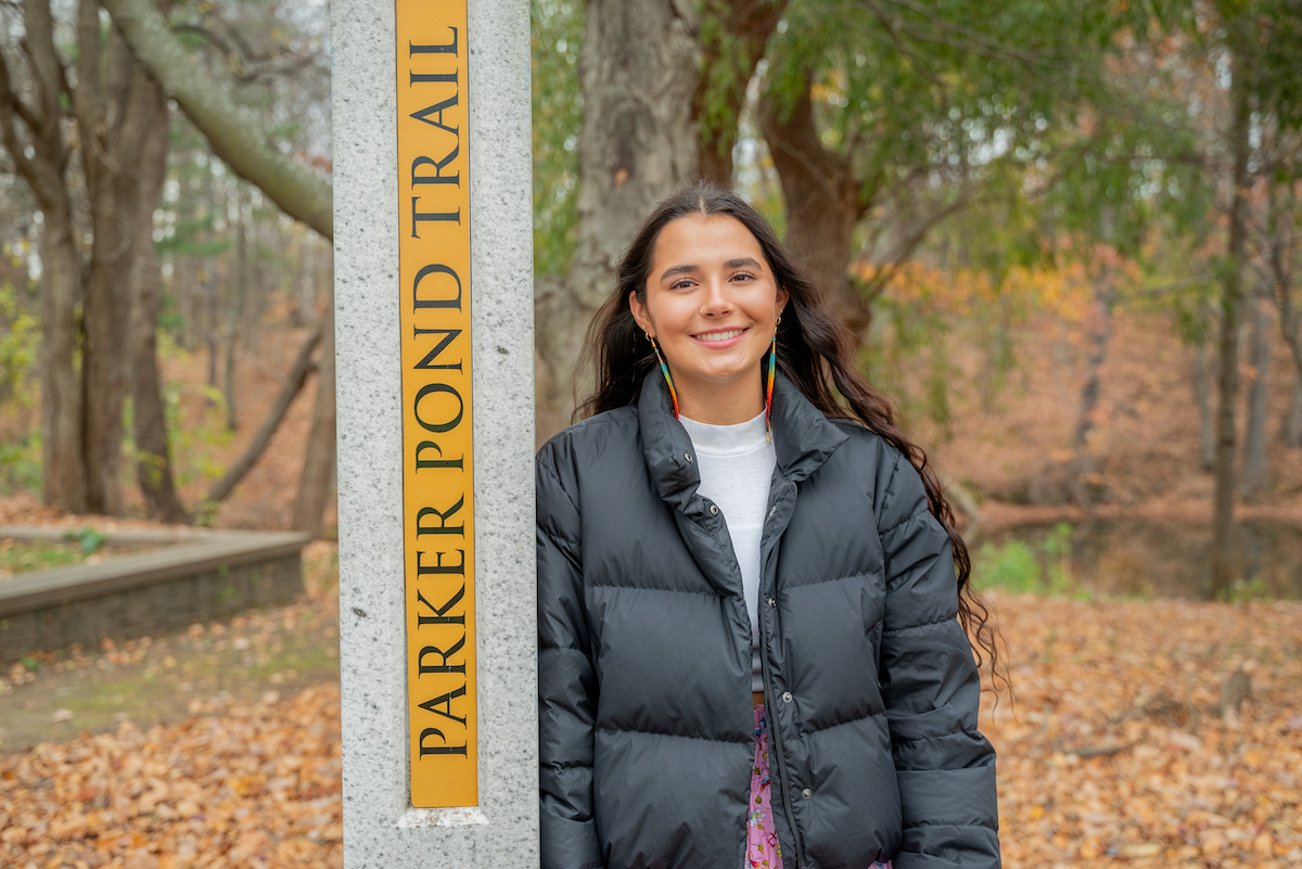 A woman with long, wavy brown hair smiles as she leans against a sign post that reads "Parker Pond Trail"