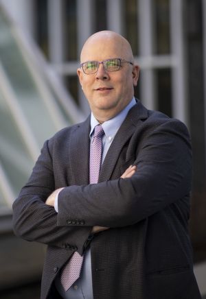 a man in glasses and a gray suit stands, arms crossed