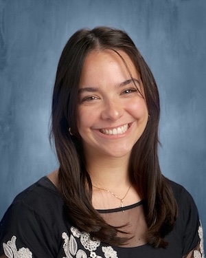 A young woman with long dark hair in a black blouse smiles for a portrait against a blue backdrop