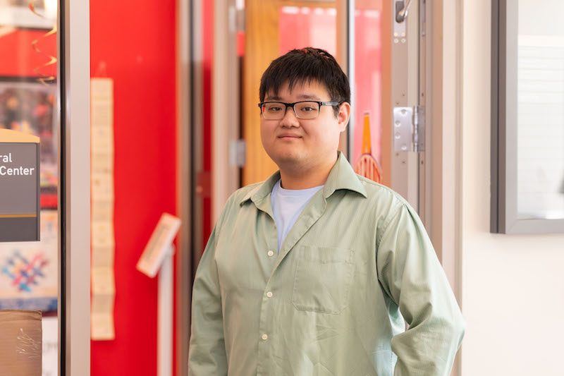 A young man with short black hair and glasses poses for a portrait against a red wall.