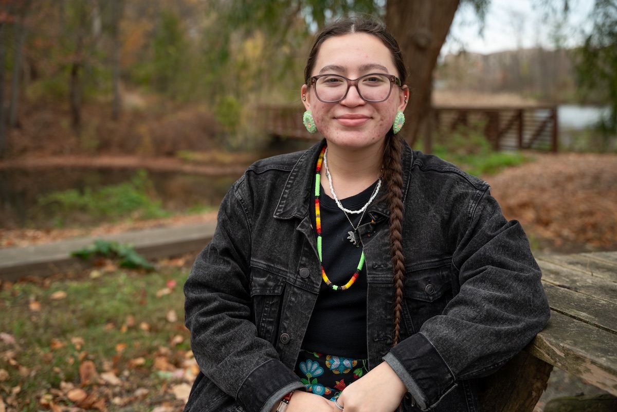 A woman wearing a long braid, glasses, black jean jacket and beaded necklace sits at a picnic table in front of a pond on an autumn day.