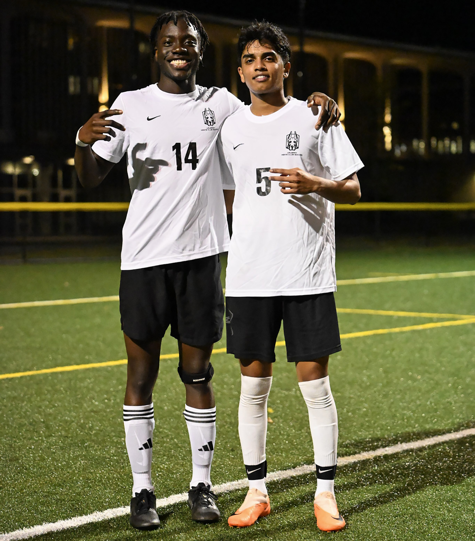 Two men in white soccer shirts and black shorts stand on a soccer pitch at night.