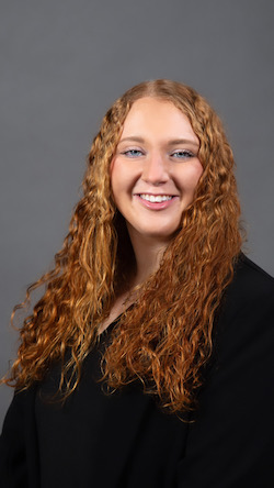 A young woman with long curly red hair wears a black shirt and smiles for a portrait against a gray backdrop. 