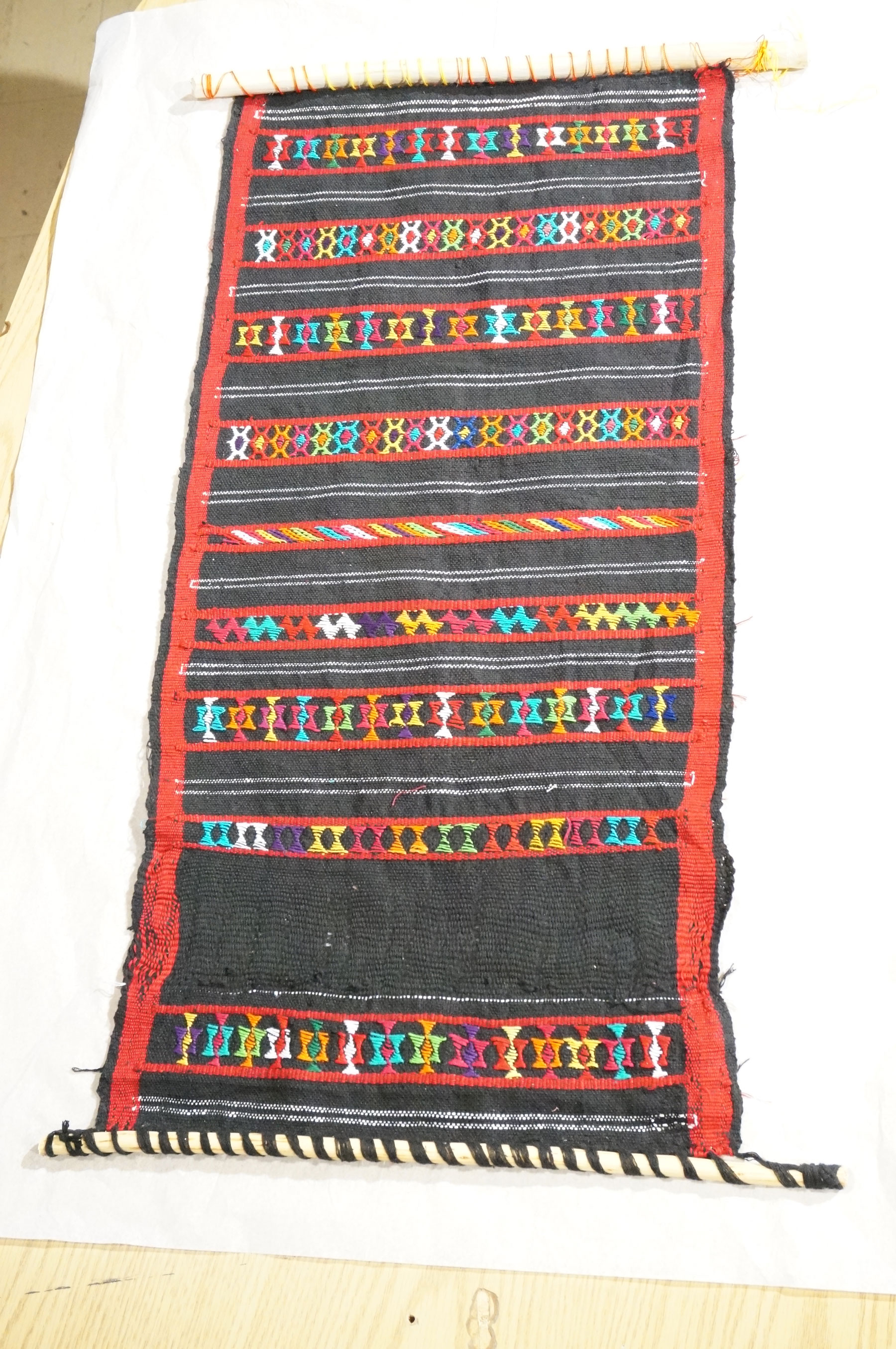 A red and black weaving with multicolored embellishments on a small loom.