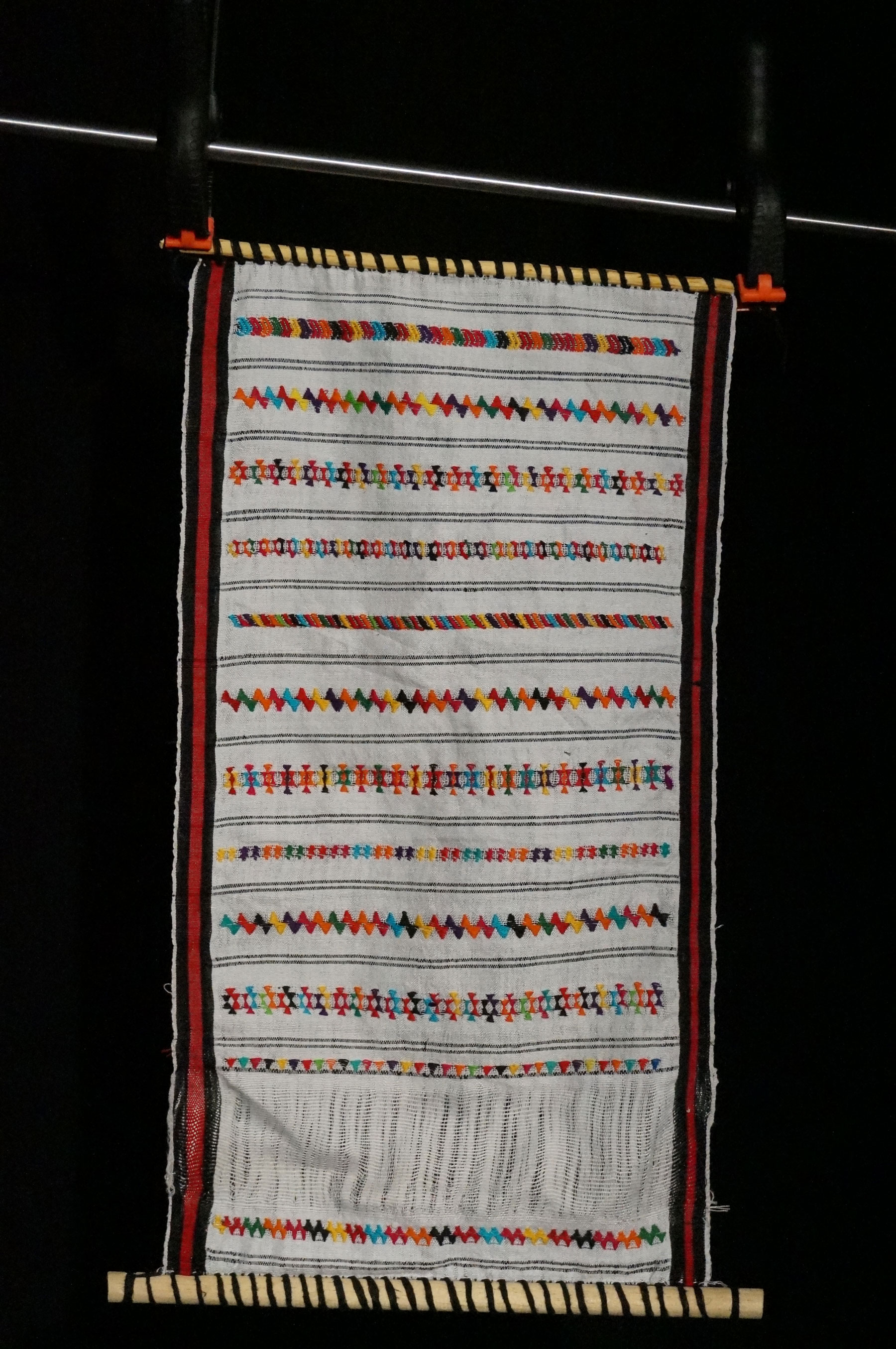 A red and white weaving with multicolored embellishments on a small loom.