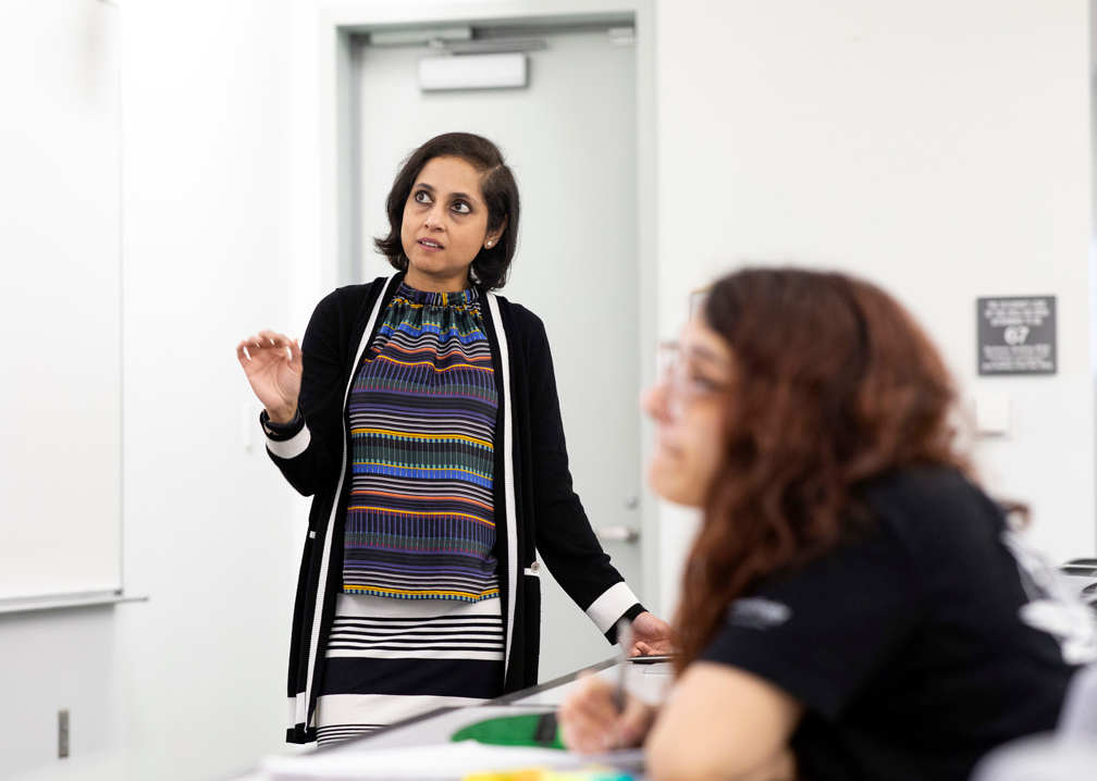 Sreya Kolay stands in front of a classroom teaching. A student sits out of focus in the foreground with long auburn hair and glasses.