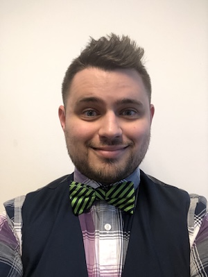 A young man with short brown hair wearing a plaid checkered shirt, dark vest and blue and green striped bowtie smiles for a portrait against a white backdrop.