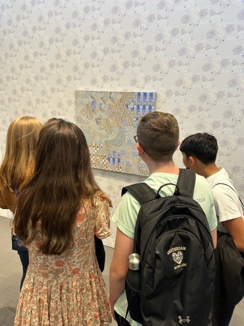 Two girls and two boys stand with their backs to the camera as they take in a piece of artwork at the University Art Museum. One wears a backpack that says "Amsterdam Bowling."
