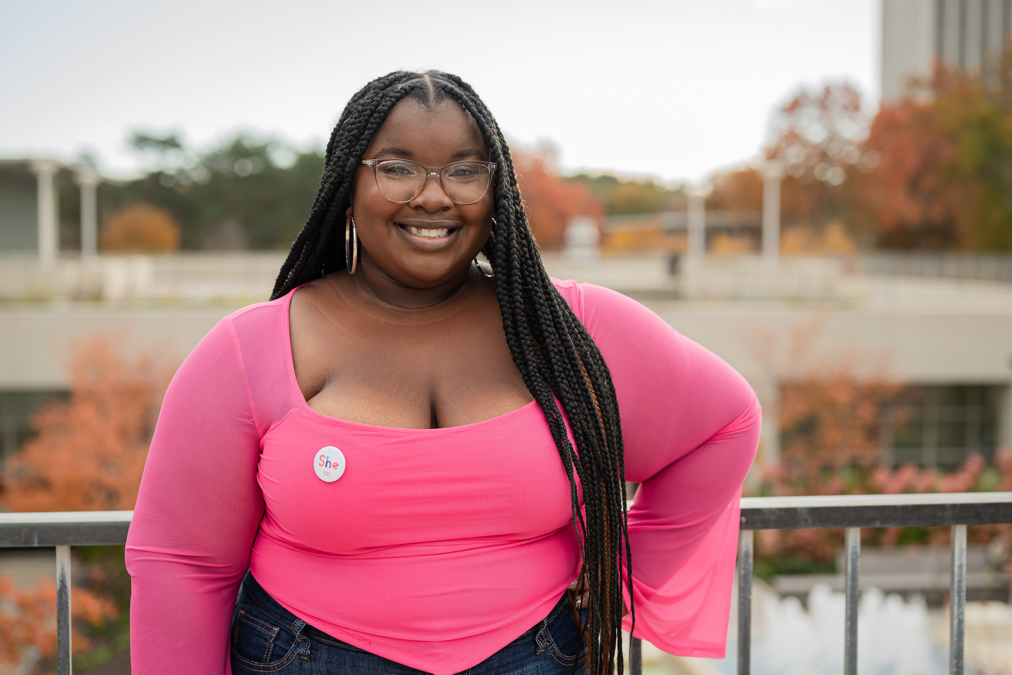 A young woman with long black hair in braids and a bright pink blouse poses on the UAlbany campus. Fall foliage can be seen in the background.