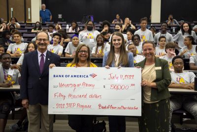four smiling people hold an enormous check for $50,000 as students in the background smile and clap