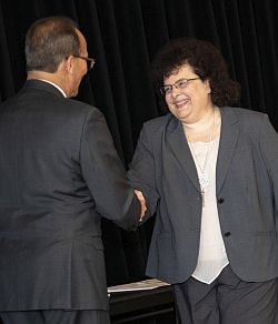President Rodriguez, his back to the camera, shakes the hand of a smiling Jeanette Altarriba during an awards ceremony.