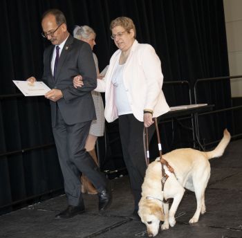 President Rodriguez walks across a stage with Delores Cimini and her white guide dog Dora