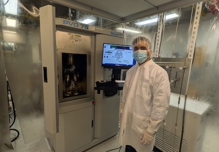 A man in a white coat, mask, hair cap and gloves stands in front of laboratory equipment.