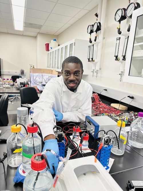 A man with short black hair and a beard in a white lab coat and blue gloves handles laboratory equipment