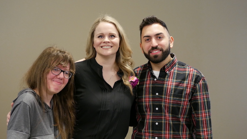 A woman with long blonde hair poses for a picture with two students, a woman with glasses and long brown hair and a man with short dark hair and a beard.