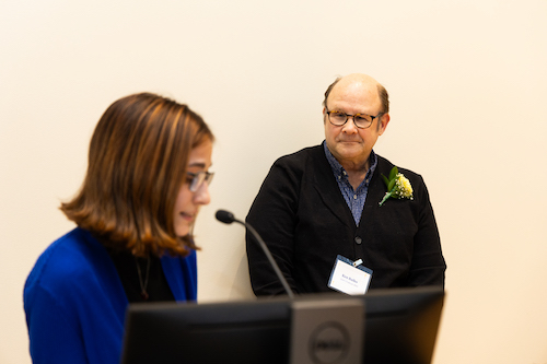 A man with glasses wears a flower lapel pinned to a sweater and leans against a wall as a student speaks into a microphone
