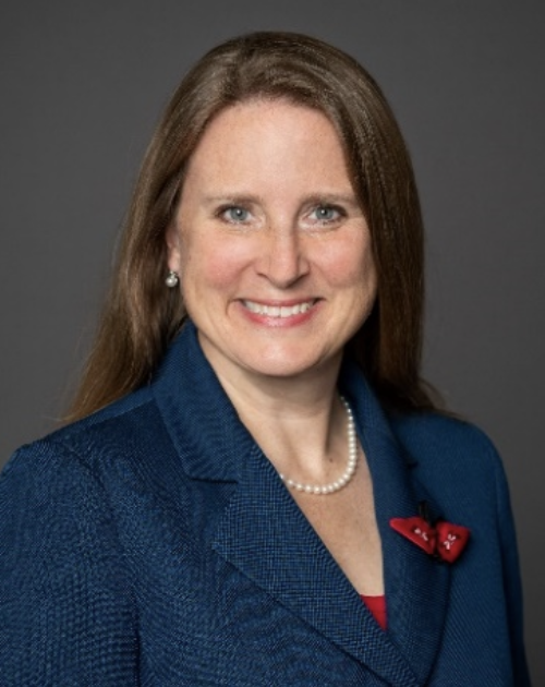 A woman with long blonde hair smiles for a portrait in a navy blazer and red brooch.