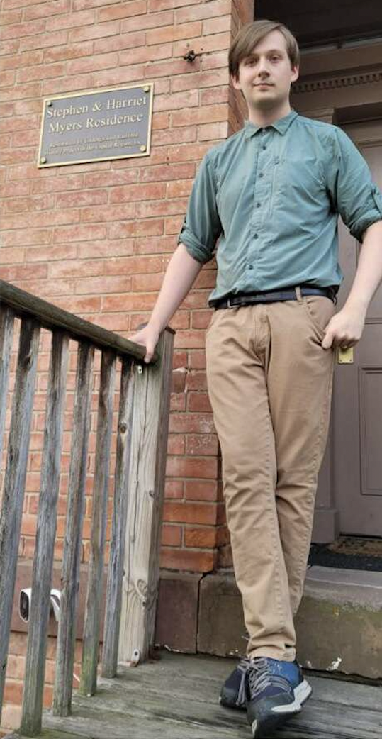 A young man with light brown hair in a button down shirt and tan pants poses in front of a brick building next to a sign that reads "Stephen and Harriet Myers Residence."