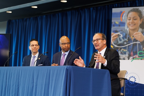 Three men in suits are seated at a table in front of microphones. One of them is speaking and gesturing.