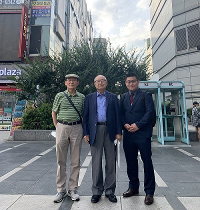 Three men pose for a photograph along a plaza in South Korea on a cloudy day.