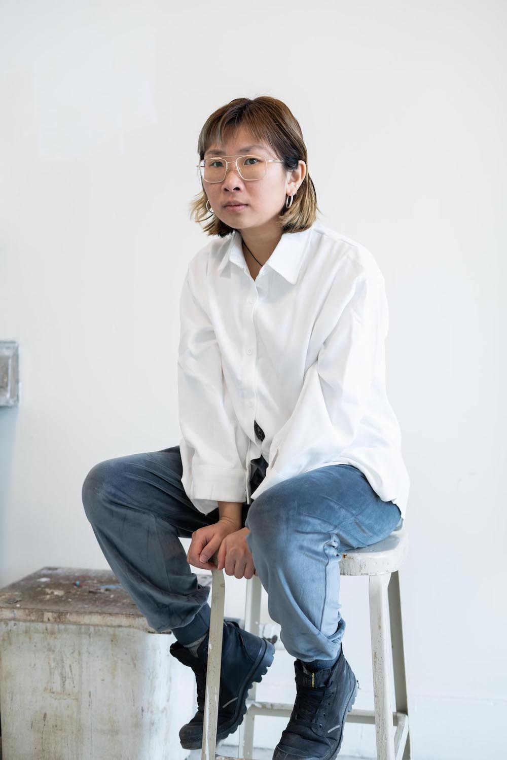 A woman with short brown hair wears a white button-down shirt and jeans and sits on a metal stool