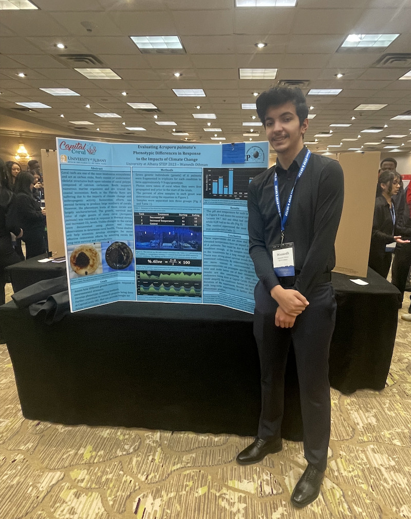 A student dressed in black dress clothes and a name tag stands, hands clasped, next to a poster board depicting coral reef research in a hotel conference room. 