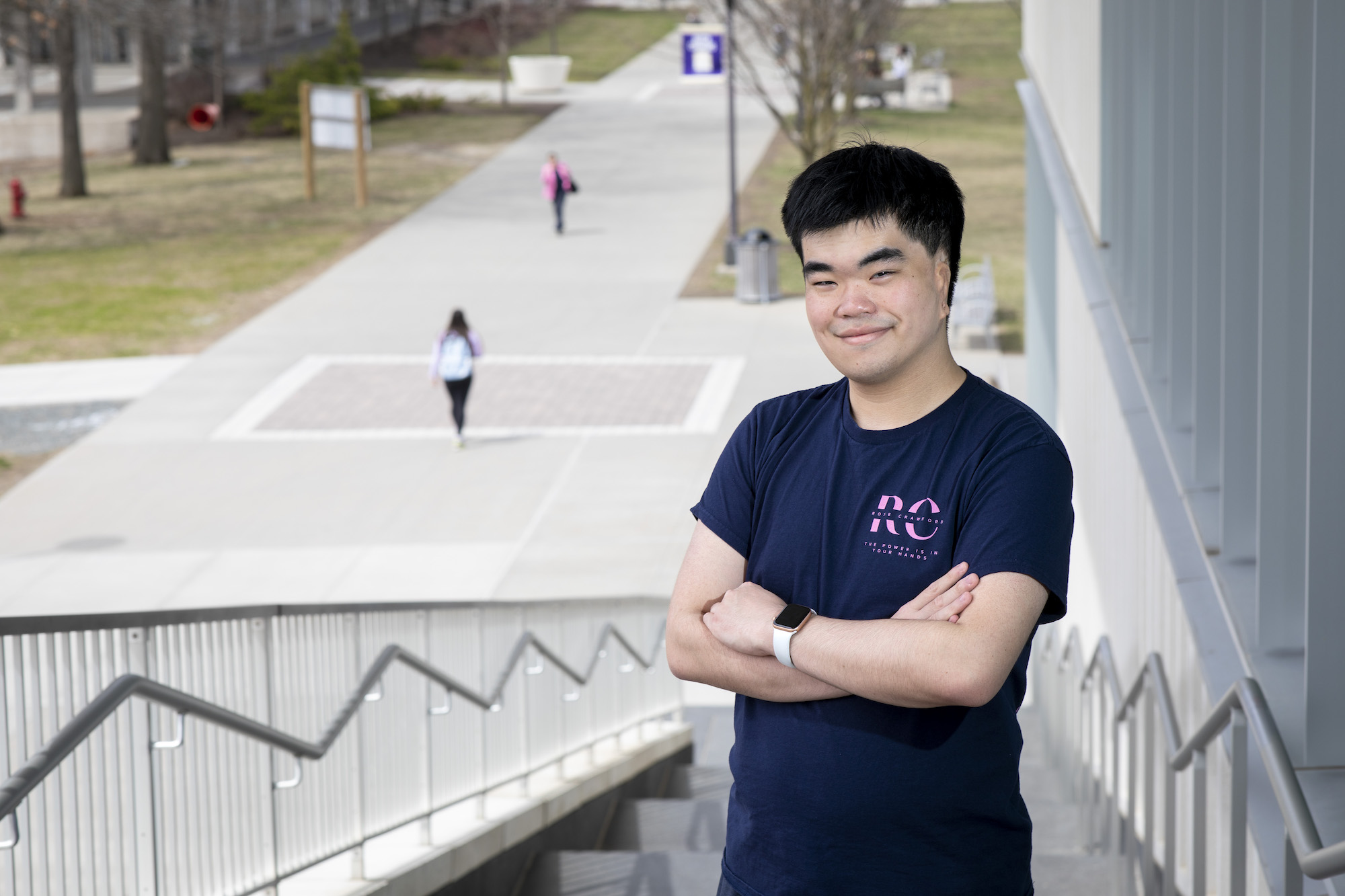 A man with short black hair smiles, arms crossed in front of his chest, as he poses for a portrait on a set of stairs on the UAlbany campus. Students can be seen walking in the background.