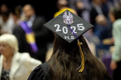 A student with long dark hair wears a cap and gown at a commencement ceremony. The cap has a Great Dane logo and reads "2025 M.S."