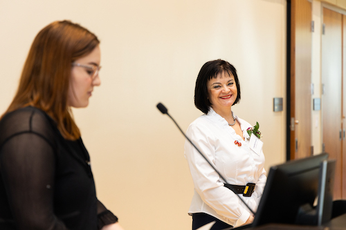 A woman with short black hair and bangs wears a white blouse and smiles as a student speaks into a microphone nearby