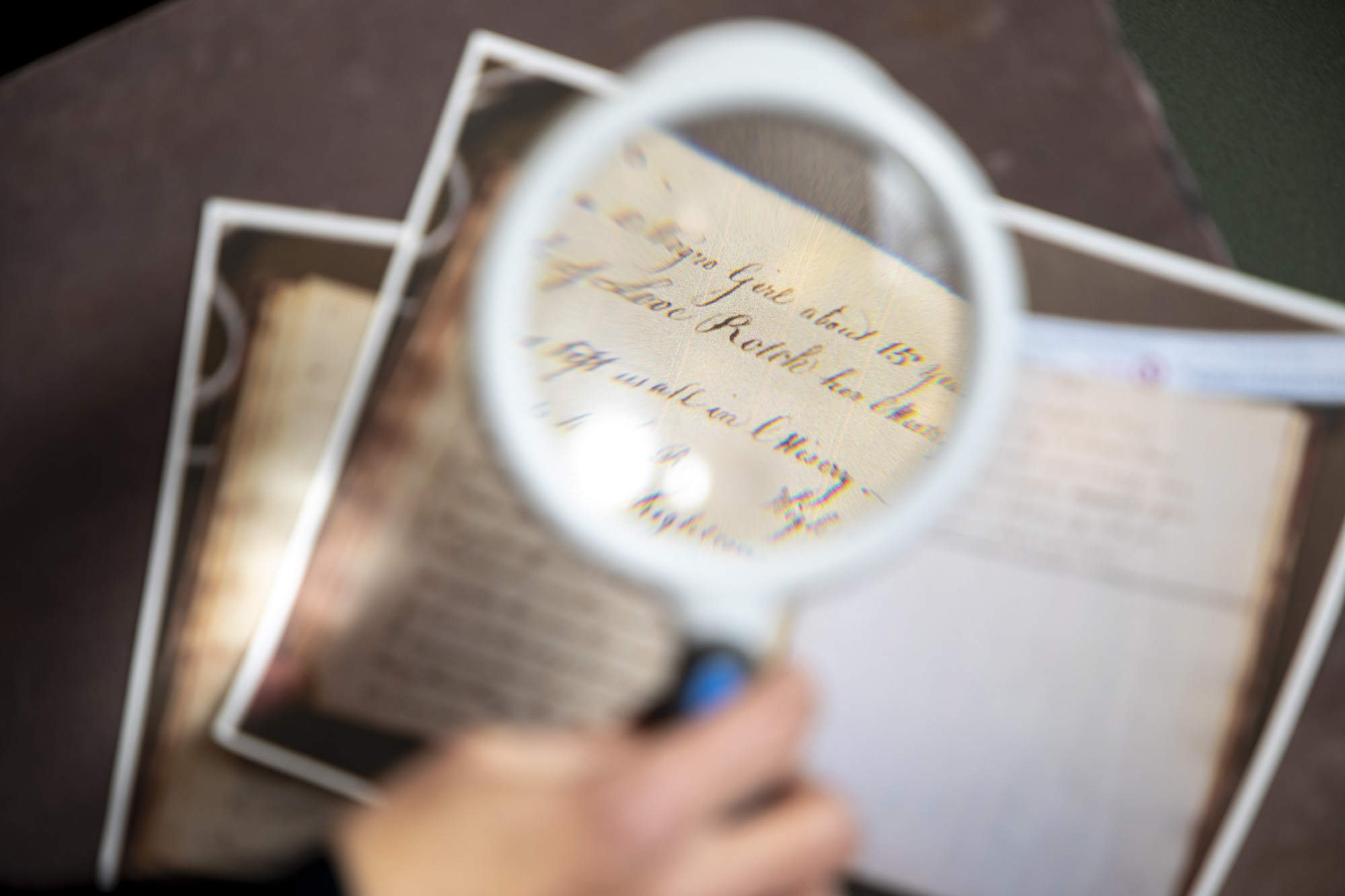 A magnifying glass is held up to a photocopy of an old poem written in elegant cursive script on a page that has been yellowed and faded from age.