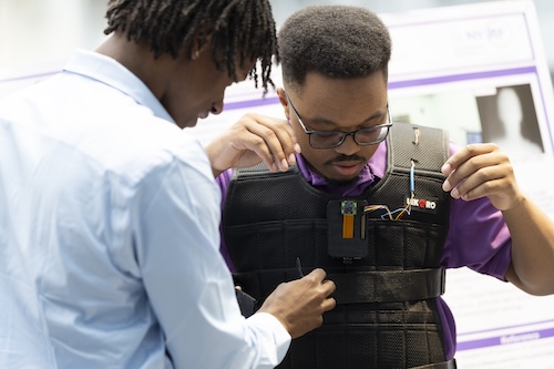 A man with short black braids in a light blue shirt adjusts a wire attached to a vest worn by a man with short black hair and glasses.