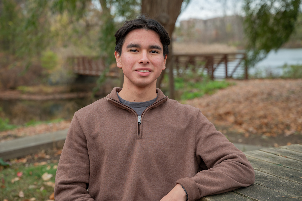 A young man with short black hair wears a brown quarter-zip sweater and sits at a picnic table in front of a pond on an autumn day.